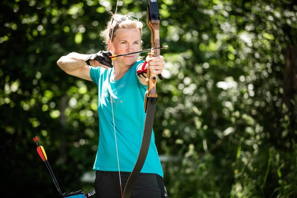 Woman Aims with a Bow in Nature, Surrounded by Green Landscape.