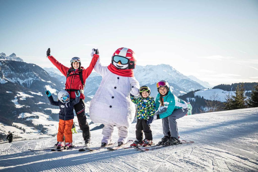 Familie beim Skifahren in den Tiroler Alpen mit einem Schneemann-Maskottchen im Hintergrund.