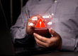 Person holding smartphone with warning symbol and notification icons in front of a laptop