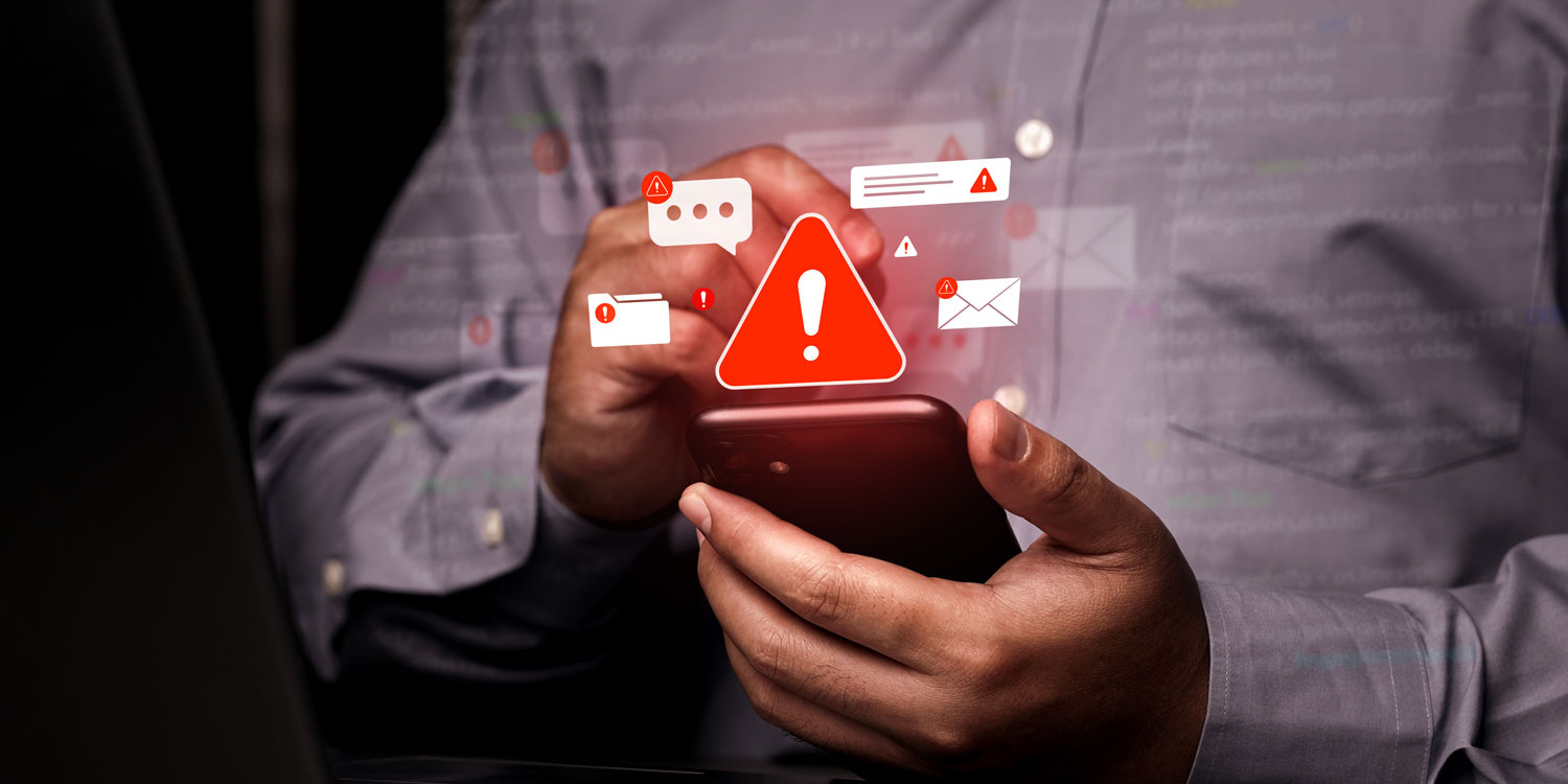 Warning About Phishing Messages Person holding smartphone with warning symbol and notification icons in front of a laptop