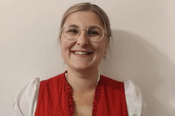 Donna in abito tradizionale rosso con gli occhiali sorride gentilmente alla fotocamera.